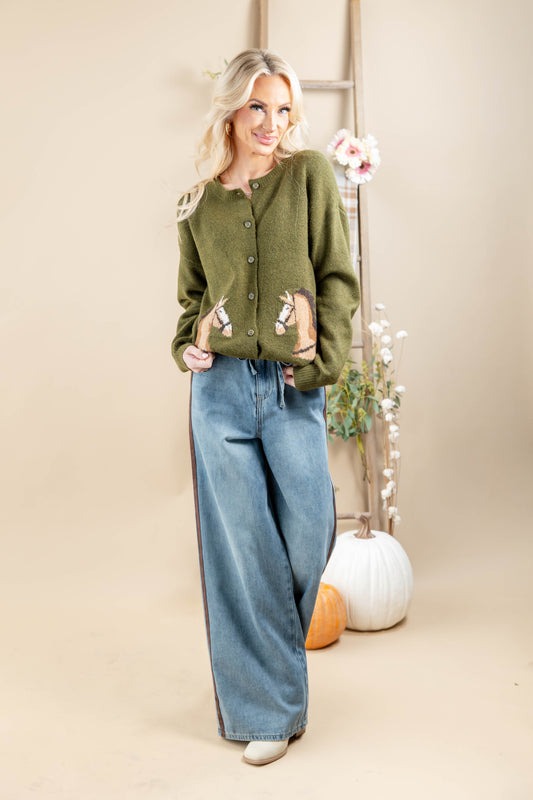 A woman modeling an olive green knit cardigan with decorative embroidered details on the pockets paired with wide-leg blue jeans, standing in a studio setting with pumpkins and a ladder backdrop.