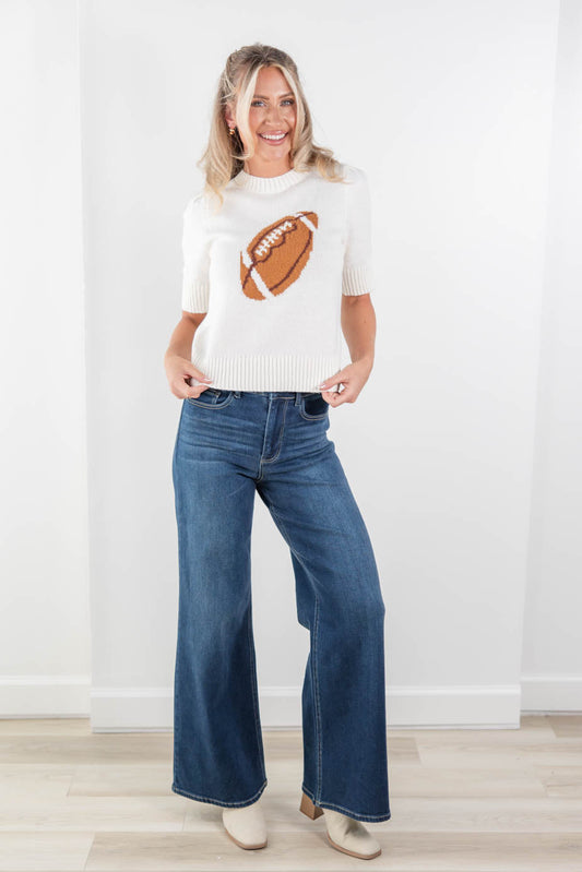 A woman stands in a bright indoor room wearing a cream knit sweater with a brown football graphic on the front, paired with wide-leg blue jeans and beige shoes.