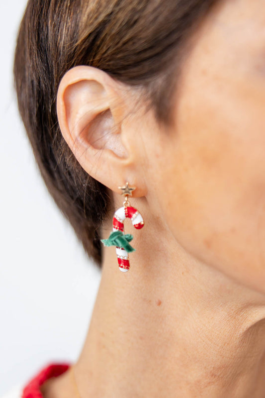 A close-up of a woman's ear wearing a dangling candy cane earring with a gold star stud, a red-and-white striped candy cane charm, and a small green holly accent.