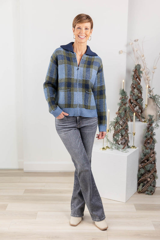 A woman modeling a blue and green plaid zip-up sweater with a navy collar, paired with gray jeans and beige boots.