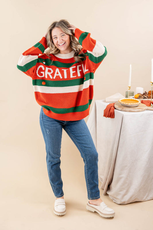 A woman modeling a festive striped sweater with 'GRATEFUL' written across the chest in white. The sweater features horizontal stripes of orange, green, and cream.