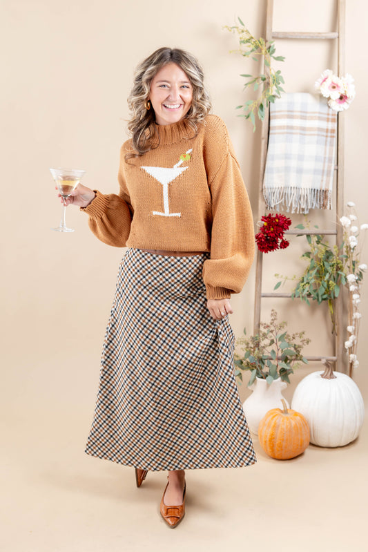 A woman stands in a styled studio setting wearing a camel-colored knit sweater with a white martini glass graphic and a black-and-beige plaid midi skirt. She holds a cocktail glass and is posed with pumpkins, a plaid throw, and floral decor against a warm beige backdrop.
