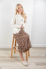 A woman sitting on a wooden stool wearing a cream-colored long-sleeve top and a brown floral midi skirt, paired with cream loafers, in a bright room with light wood floors.