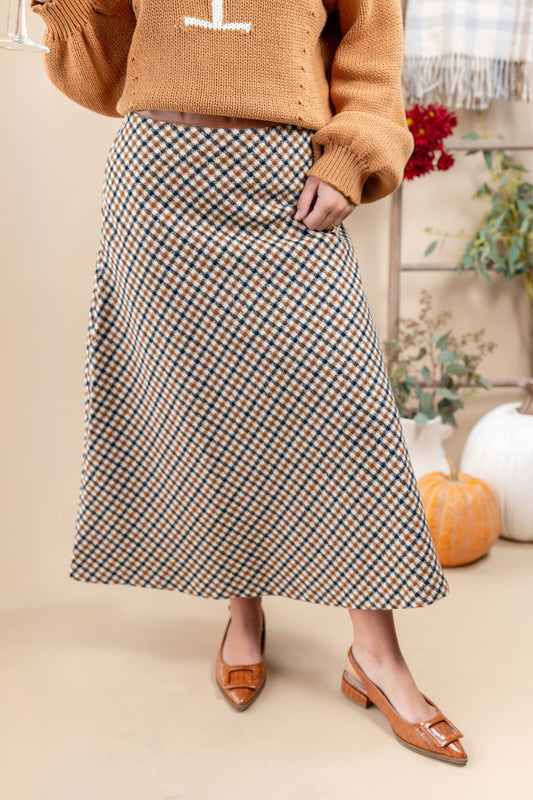 A woman wearing a long A-line skirt with a brown, beige, and white gingham pattern, styled with a tan knit sweater and tan pointed-toe slingback flats. The background is a warm beige studio setting with decorative pumpkins and a ladder.