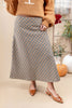 A woman wearing a long A-line skirt with a brown, beige, and white gingham pattern, styled with a tan knit sweater and tan pointed-toe slingback flats. The background is a warm beige studio setting with decorative pumpkins and a ladder.