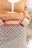 Close-up of a woman wearing a tan knit sweater and a brown-and-gray plaid skirt, pulling a tan faux leather waistband outward with both hands.
