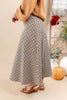 A person wearing a long pleated skirt with a beige, brown, and black gingham pattern, paired with a tan sweater and brown flats, standing in a styled indoor setting with a pumpkin and plants.