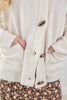 Close-up of a cream-colored knit cardigan sweater with brown wooden buttons and side pockets, worn by a model.
