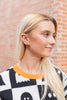 A blonde woman wearing a black and white checkered sweater with pumpkin jack-o-lantern stud earrings, standing against a brick wall.