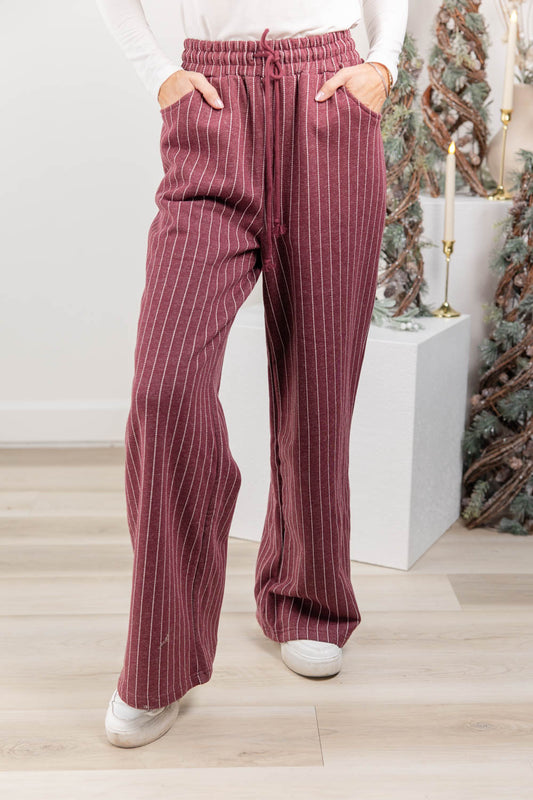 A woman wearing burgundy wide-leg pinstripe pants with a drawstring waist, paired with a white long-sleeve top and white sneakers.