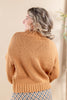 A woman shown from the back wearing a warm camel-colored knit sweater with textured cable and baffle details, paired with a brown and white checkered skirt, set against a soft beige background.