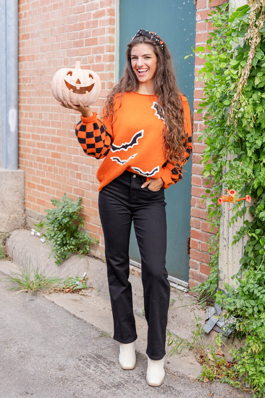 A woman stands outdoors in a brick alleyway wearing a bright orange Halloween sweater with black and orange checkered sleeves, black bat motifs, and black-and-white striped trim. She is also wearing black pants and white ankle boots, and holds a carved jack-o'-lantern pumpkin.