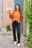 A woman stands outdoors in a brick alleyway wearing a bright orange Halloween sweater with black and orange checkered sleeves, black bat motifs, and black-and-white striped trim. She is also wearing black pants and white ankle boots, and holds a carved jack-o'-lantern pumpkin.