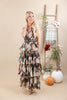 A woman modeling a sleeveless, tiered maxi dress with a brown, beige, and teal floral pattern, standing in a studio decorated with a wooden ladder, pumpkins, and a plaid throw blanket.
