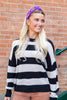A woman with long blonde hair wearing a black and white striped knit sweater, brown pants, and a purple halloween patterned headband, standing in front of a red brick wall.