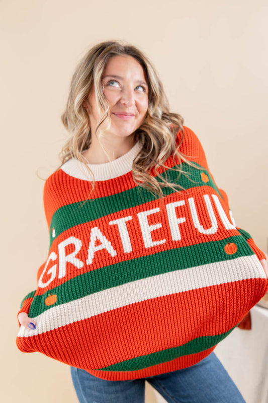 A woman wearing a festive, oversized knit sweater with horizontal stripes of orange, green, and white, featuring the word 'GRATEFUL' in white letters across the chest. She is posing against a solid beige background.