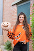 A woman stands outdoors wearing a bright orange Halloween sweater with black and orange checkered sleeves, black bat motifs, and black-and-white striped trim and holds a carved jack-o'-lantern pumpkin.