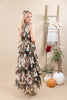 A woman stands in a studio wearing a sleeveless, tiered maxi dress with a floral print in brown, beige, and muted teal tones. The background is a warm beige with a wooden ladder decorated with a plaid throw, flowers, and pumpkins.
