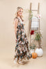 A woman models a sleeveless, tiered maxi dress with a brown, beige, and teal floral pattern. She is standing in a studio setting with a beige backdrop, next to a wooden ladder decorated with a plaid throw blanket, flowers, and pumpkins.