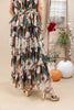 A woman wearing a brown tiered maxi dress with a floral print in pink, white, and teal, paired with gold heeled sandals, standing in a styled indoor setting with pumpkins and a ladder.