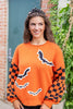A woman wearing a bright orange crewneck sweater with black and orange checkered sleeves and black bat motifs, standing outdoors in front of a brick wall and greenery.