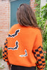 A woman from behind wearing a bright orange Halloween sweater with black and orange checkered sleeves, black bat motifs, and black-and-white striped trim. She is also wearing black pants.