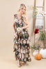 A woman models a sleeveless, tiered maxi dress with a floral print in earthy tones of brown, beige, and teal. She is standing in a styled studio setting with a wooden ladder, a plaid throw blanket, and decorative pumpkins and vases.