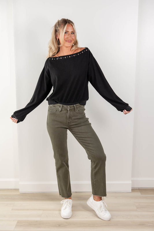 A woman modeling a black off-the-shoulder long-sleeve top with silver stud embellishments, paired with olive green jeans and white sneakers, standing against a white wall.