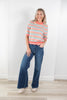 A woman modeling a short-sleeve knit sweater with horizontal pastel stripes in pink, orange, and blue, paired with dark blue wide-leg jeans and beige sneakers, standing against a white wall.