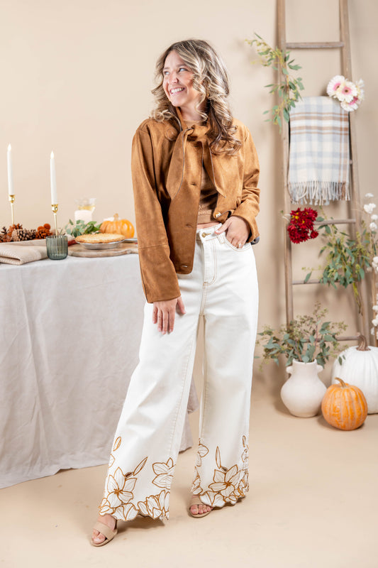 A woman stands in a styled indoor setting wearing a brown suede-like button-front jacket over a tan top and white wide-leg pants with gold floral embroidery near the hems. The background includes a table with candles and autumn decor, a ladder with a plaid throw, and pumpkins.