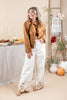 A woman stands in a styled indoor setting wearing a brown suede-like button-front jacket over a tan top and white wide-leg pants with gold floral embroidery near the hems. The background includes a table with candles and autumn decor, a ladder with a plaid throw, and pumpkins.