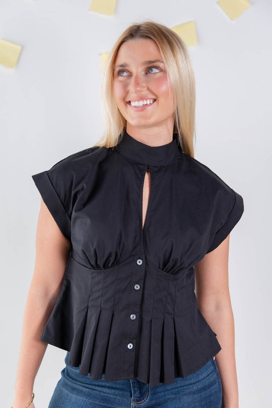 A woman wearing a black sleeveless mock-neck blouse with a keyhole cutout, gathered waist, and pleated peplum hem, paired with blue jeans against a light gray wall.