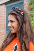 A woman with long brown hair wearing a black fabric headband decorated with colorful patches, beads, and small metal studs, photographed outdoors in front of a brick wall and greenery.
