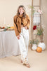 A woman modeling a brown suede-like jacket and white wide-leg pants with gold floral embroidery near the hem, standing in a styled indoor autumn setting with pumpkins, a table, and a ladder shelf.