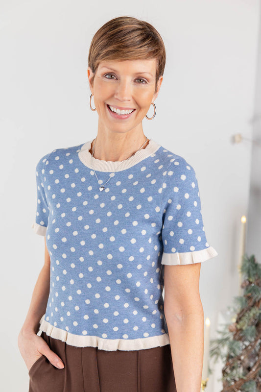 A woman wearing a light blue short-sleeve knit top with white polka dots and cream ruffle trim at the collar, cuffs, and hem, paired with brown pants.