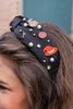 A close-up of a woman wearing a black fabric headband decorated with Halloween-themed enamel pins, including jack-o'-lanterns, a witch hat, and faux pearls, with her brown hair visible.