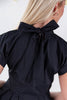 Close-up back view of a woman wearing a black short-sleeve top with a large bow detail at the back of the neck.