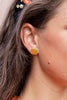 A close-up of a woman's ear wearing a small pumpkin shaped stud earring with a bright orange center and a gold-toned outer ring.
