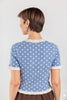 A woman shown from the back wearing a light blue short-sleeve top with white polka dots and white ruffled trim at the collar, cuffs, and hem.