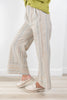 A person wearing loose, wide-leg striped pants in beige, cream, and muted blue tones, paired with white slip-on shoes and a lime green top, standing on a light wood floor against a white wall.