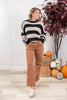 A woman modeling a black and beige striped knit sweater and brown wide-leg jeans, standing in a decorated indoor room with pumpkins and a potted plant.