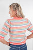 A woman shown from the back wearing a short-sleeve knit sweater with horizontal stripes in coral pink, blue, white, and light green, paired with blue jeans against a plain light gray background.