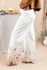 White wide-leg pants with gold floral embroidery near the hem, shown on a model wearing a brown jacket and sandals in an indoor setting.