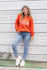 A woman wearing a bright orange crewneck sweater with the word 'Boo' written across the front in large white cursive letters, paired with blue jeans and white sneakers.