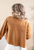 A woman viewed from behind wearing a cropped, long-sleeve brown suede or faux-suede jacket over white pants, set against a soft beige indoor background.