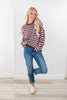 A woman modeling a maroon and white horizontally striped knit sweater, paired with blue jeans and white slip-on shoes, standing against a white wall and light wood floor.