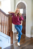 A woman posing on a staircase landing wearing a burgundy short-sleeve button-up blouse with white polka dots, blue jeans, and white loafers.