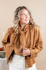 A woman wearing a camel brown suede-like jacket with a large matching bow tie detail, styled over a brown top and white pants, posing against a warm beige background.