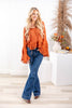 A woman wearing a rust orange long-sleeve blouse with a V-neck, ruffle hem, and flared cuffs paired with jeans.