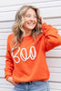 A woman wearing a bright orange crewneck sweater with the word 'Boo' written across the front in large white cursive letters, paired with blue jeans.